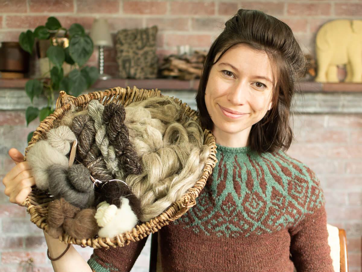 Marina is wearing a colourwork yoke jumper and holding a basket of naturally coloured fibres for spinning