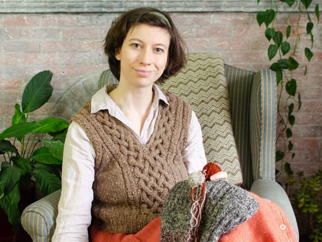 Marina sits in a comfy chair in a hand-knitted cabled tank top, with a knitting project and yarn on her lap
