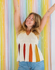 A woman in a colorful knit top and high-waisted jeans poses joyfully against a backdrop of colourful streamers. The knitted top is white with a peplum bottom which has colourful folds.