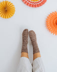 A close up shot of a persons legs held up against a wall. They are wearing white trousers and a speckled pair of handknit socks. The wall is adorned with colourful paper decorations.