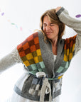 A woman in a white dress wears a grey handknitted cardigan that features colourful checkered colourwork in two panels on the front.