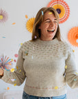 A woman in a handknitted sweater with cute confetti like detailing across the yoke stands amidst colorful confetti, with festive paper decorations in the background, celebrating joyfully.