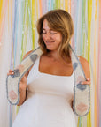 A woman in a white dress wraps a short, hand knitted scarf around the back of her neck. She is holding each end and looking down smiling. Behind her is a backdrop of pastel streamers.