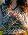 a torso shot of marina. She is wearing a colourwork handknit vest with branch like motifs, but the focus is on her hands held in front of her chest in a pair of colourwork mitts knit in sheepy shades.