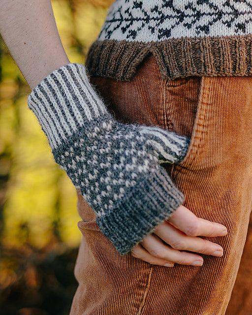 A close up image of a hand knitted wristwarmer made using grey and white undyed British Wool. The mitt has a 2 colour ribbing at the wrist and thumb, a 2 colour stranded colourwork design on the hand and a folded brim at the fingers.