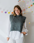 A person wears a gray hand knitted vest with floral embroidered detailing over a white shirt. She is smiling directly at the camera and there is colourful confetti and circular decorations in the background.