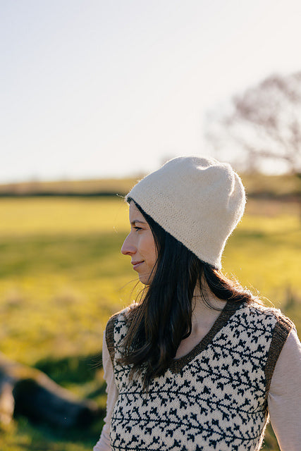 Marina, a white woman with brown hair is standing outside and pictured looking to the side. She is wearing a handknit colourwork vest, but the focus is on the white felted knit hat she is wearing.