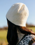 The back view of Marina, a white woman, wearing a creamy white hand-knit hat with a subtle herringbone texture around the brim. Blue sky and countryside fields are blurred in the background.