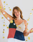 A woman in a colorful, patchwork knit top raises her arm, surrounded by gold and white decorative circles hanging on a white wall.