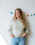 A woman with long, wavy hair wears a cosy light hand knitted sweater and high-waisted jeans. She is posing against a wall decorated with colorful dots and smiling at someone just off centre.