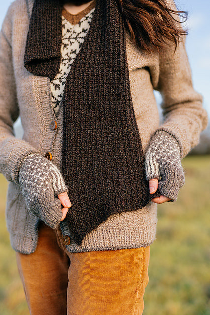 A body shot of Marina wearing a cosy grey cardigan, fingerless mitts and a  dark brown handknit scarf. She is holding the ends of the scarf so that you can see the gentle taper.