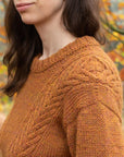 A close up shot of the shoulder of a person wearing a a rust-colored, textured handknit sweater. The focus is on the details, in particular the drop shoulder and knitted cables.