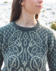 A close-up of Marina, a dark haired white woman wearing a green hand-knitted sweater with detailed botanical colourwork on the chest. The snowy background is softly blurred making the jumper the focus of the image.