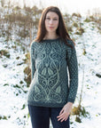 Marina, a dark haired, white woman stands facing the camera in a snowy outdoor setting, wearing a green hand-knitted sweater with an intricate botanical inspired colourwork design on the front. The sweater is paired with dark trousers, and the natural surroundings of snow-covered hills and sparse trees add a wintry charm to the scene.