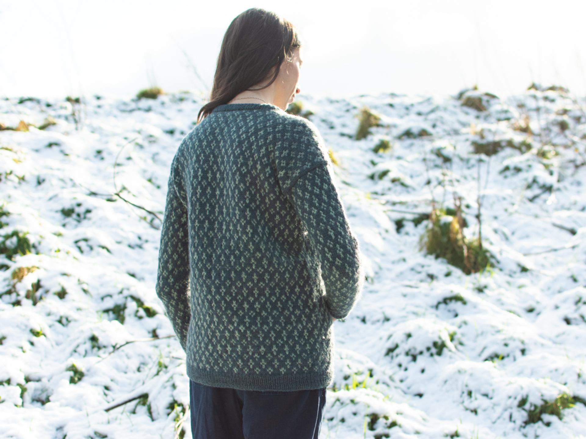 Marina, a dark haired white woman is pictured from behind standing outdoors in a snowy landscape. She is wearing a green hand-knit sweater, with her hair flowing down her back. The sweater features a delicate, repeating design and is paired with dark trousers. The snowy hill and bright sky create a serene winter backdrop.