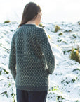 Marina, a dark haired white woman is pictured from behind standing outdoors in a snowy landscape. She is wearing a green hand-knit sweater, with her hair flowing down her back. The sweater features a delicate, repeating design and is paired with dark trousers. The snowy hill and bright sky create a serene winter backdrop.