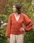 A front facing shot of Marina Skua's Amber Apple cardigan. Marina is stood with a hand on her hip, looking to the side. The cardigan is orange with a v neck and voluminous balloon sleeves