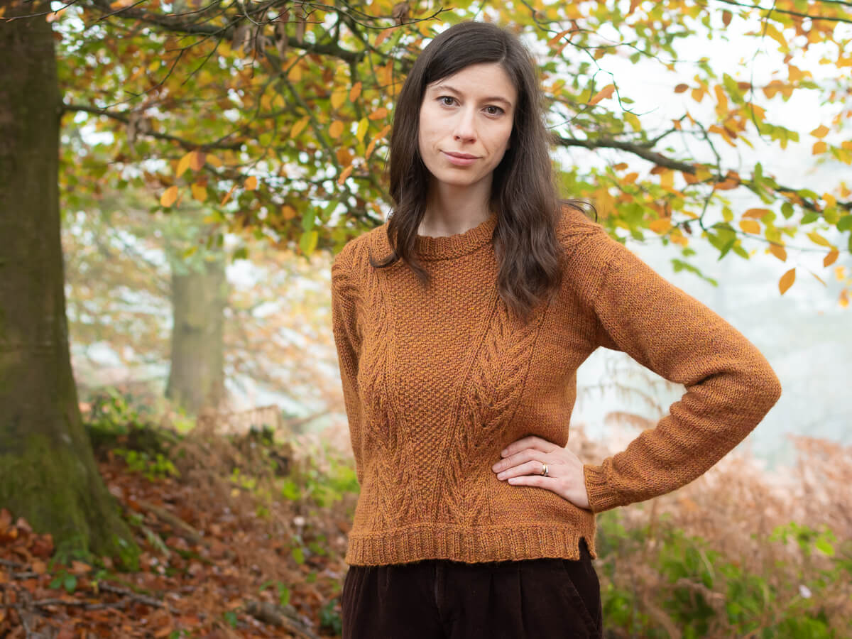 Marina, a white person with long brown hair is standing outdoors in an autumnal forest setting. She is wearing a rust-coloured, textured handknit sweater with a subtle leaf pattern, blending harmoniously with the warm, earthy tones of the fallen leaves and surrounding trees.