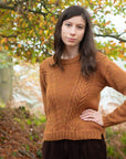 Marina, a white person with long brown hair is standing outdoors in an autumnal forest setting. She is wearing a rust-coloured, textured handknit sweater with a subtle leaf pattern, blending harmoniously with the warm, earthy tones of the fallen leaves and surrounding trees.