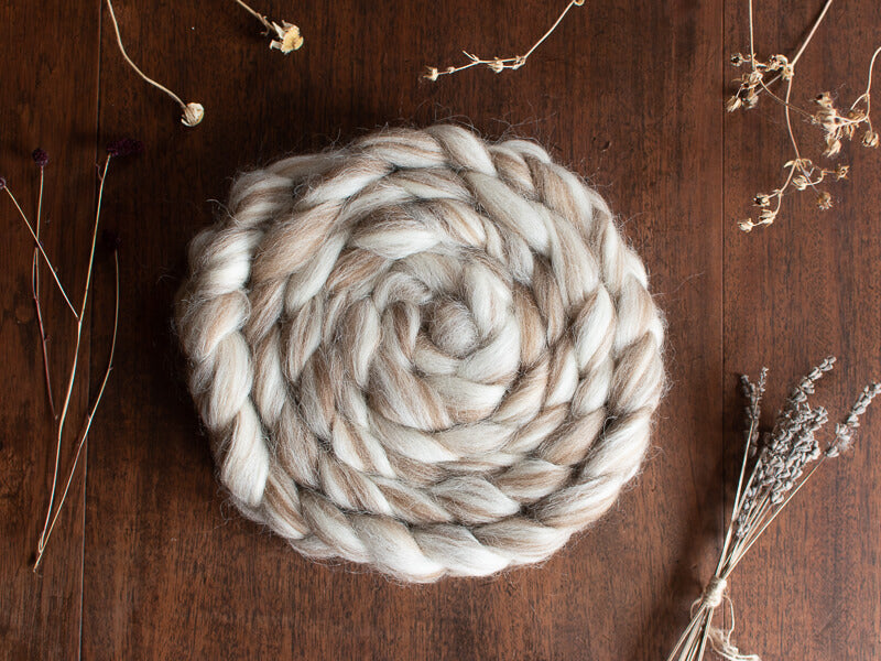 A braided bundle of cream and light brown British wool fibre resting on a wooden surface, surrounded by dried botanicals such as lavender stems and seed heads