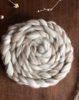 A braided bundle of cream and light brown British wool fibre resting on a wooden surface, surrounded by dried botanicals such as lavender stems and seed heads