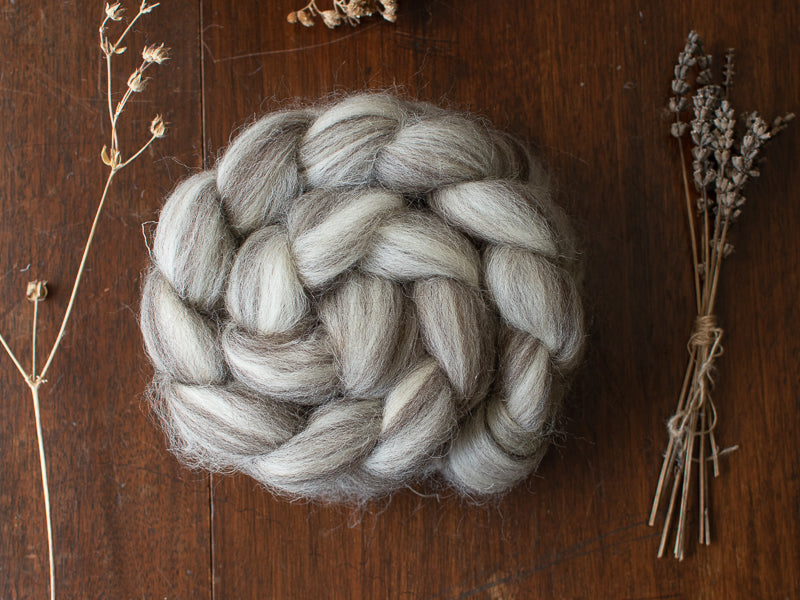 A braided bundle of light grey and grey British wool spinning fibre resting on a wooden surface, surrounded by dried botanicals such as lavender stems and seed heads
