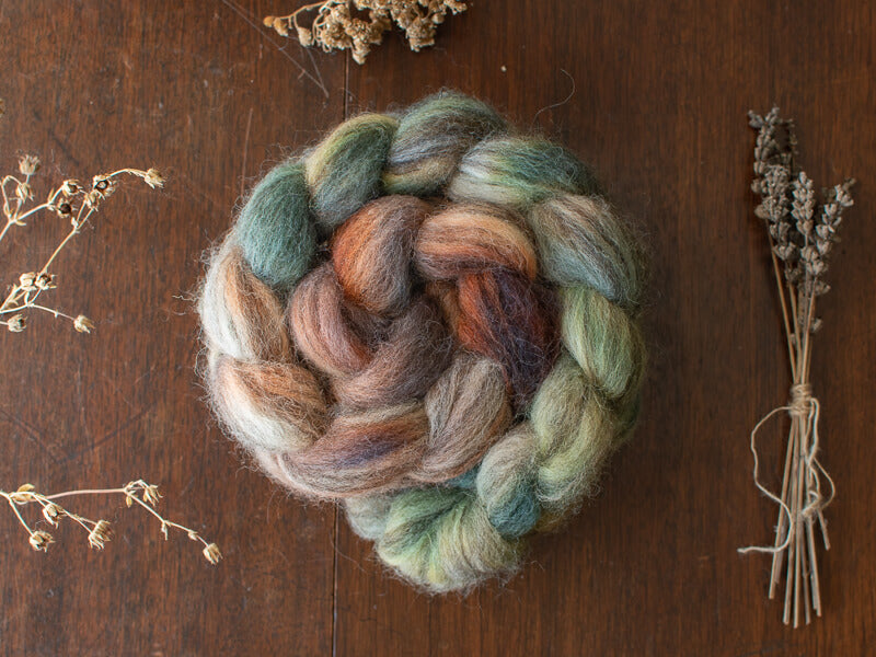 A rolled braid of hand-dyed British wool spinning fiber in earthy, muted tones, featuring colors like teal blue, , soft browns, dusky purples, and mellow greens
