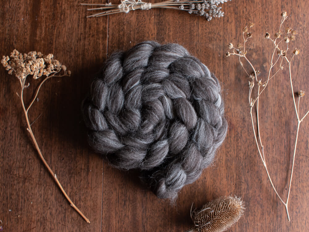 A braided bundle of dark gray British wool fibre resting on a wooden surface, surrounded by dried botanicals such as lavender stems and seed heads