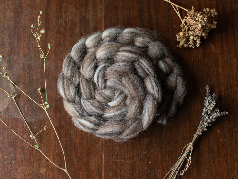 A braided bundle of grey, brown and cream British wool spinning fibre resting on a wooden surface, surrounded by dried botanicals such as lavender stems and seed heads