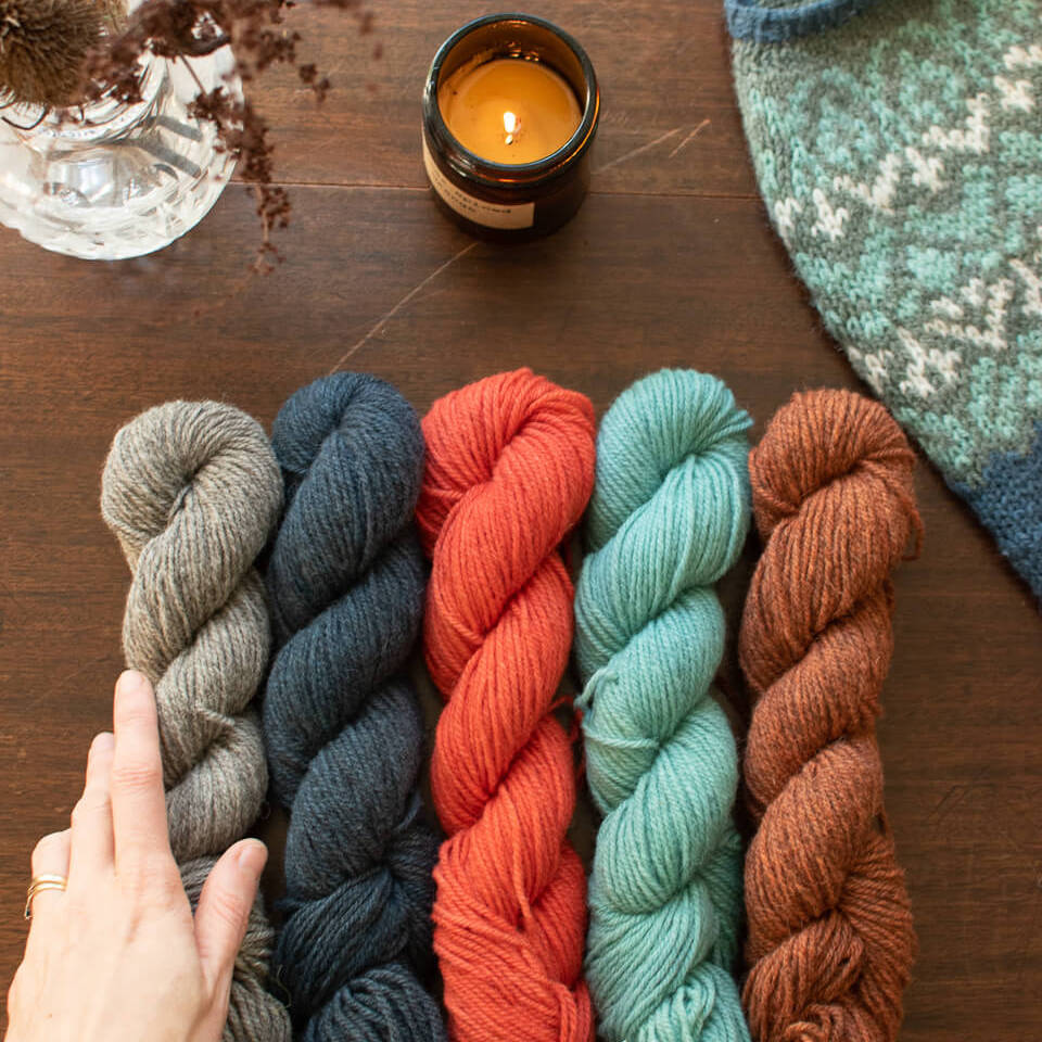 Hand-dyed yarn skeins in rich autumn shades of brown, red, orange, blue, grey. Alongside is a sliver of a hand knit colourwork yoke jumper and a lit candle
