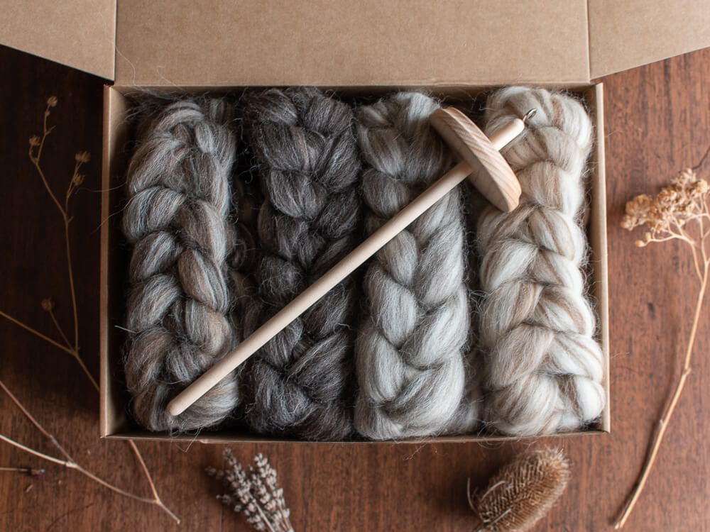 A cardboard box containing four neatly braided bundles of British wool spinning fibre in varying shades of gray and cream, displayed on a wooden surface. Dried botanicals, including seed heads and lavender, are arranged around the box, complementing the natural tones and textures of the wool. A spindle sits top the braided fibres.