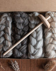A cardboard box containing four neatly braided bundles of British wool spinning fibre in varying shades of gray and cream, displayed on a wooden surface. Dried botanicals, including seed heads and lavender, are arranged around the box, complementing the natural tones and textures of the wool. A spindle sits top the braided fibres.