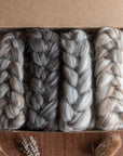 A cardboard box containing four neatly braided bundles of British wool spinning fibre in varying shades of gray and cream, displayed on a wooden surface. Dried botanicals, including seed heads and lavender, are arranged around the box, complementing the natural tones and textures of the wool.