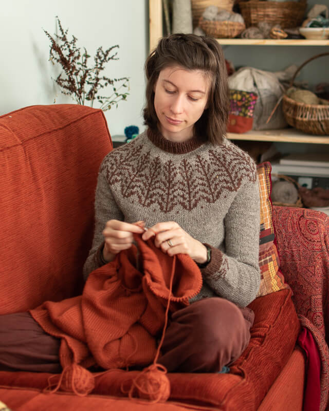 Marina is sitting knitting with orange yarn on an orange sofa, wearing a hand-knitted jumper