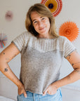 A woman poses in a light, hand knitted top with lace detail, against a backdrop of colorful paper decorations.