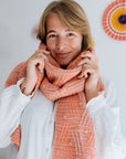 A person wearing a soft, patterned hand knit coral scarf over a white blouse, with decorative paper fans in the background.