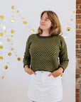 A white person in a green checkered handknit sweater made using hand dyed british wool yarn and white skirt stands smiling in front of a wall with gold decorations.