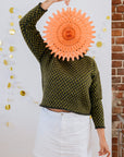 A white person in a green checkered handknit sweater and white skirt is posing for the camera holding a pink paper decoration in front of her face. On the wall behind her are some gold and white paper decorations.