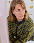 A white person in a green checkered sweater and white skirt sits looking away from the camera. Behind her is a wall with gold decorations.