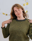 A white person in a green checkered handknit sweater and white skirt is posing and smiling for the camera with her eyes closed and hands on her shoulders. On the wall behind her are some gold and white paper decorations.