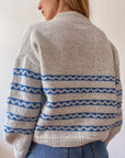 Sophie, a white woman with blonde hair is modelling her handknitted Sennen sweater. The sweater is a creamy colour with a series of blue stripes that have creamy waves in the middle of each. It features a crew neck and balloon sleeves that taper neatly just before the cuff. Sophie is stood with her back to the camera so we have a clear picture of the back of the sweater.