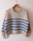 A simple product shot of a handknitted sweater. The Sennen sweater is a creamy colour with a series of blue stripes that have creamy waves in the middle of each. It features a crew neck and balloon sleeves that taper neatly just before the cuff. It is sitting on a wooden hanger, hung on a nail in the wall.