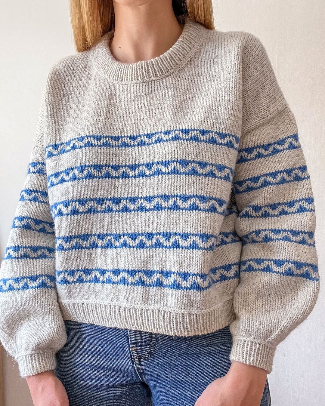 Sophie, a white woman with blonde hair is modelling her handknitted Sennen sweater. The sweater is a creamy colour with a series of blue stripes that have creamy waves in the middle of each. It features a crew neck and balloon sleeves that taper neatly just before the cuff.  Sophie is stood in front of the camera so we have a clear picture of the front of the sweater, but her face is out of shot.