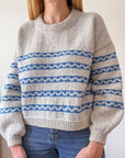 Sophie, a white woman with blonde hair is modelling her handknitted Sennen sweater. The sweater is a creamy colour with a series of blue stripes that have creamy waves in the middle of each. It features a crew neck and balloon sleeves that taper neatly just before the cuff. Sophie is stood in front of the camera so we have a clear picture of the front of the sweater, but her face is out of shot.