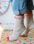 Close up shot of a persons ankles. They are wearing pastel coloured hand knitted socks with pink accents and blue rolled-up jeans, standing on a wooden floor scattered with colourful confetti.
