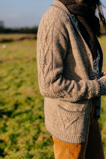 A torso shot of marina standing in a field outside. She is wearing a textured handknit grey cardigan and a dark brown scarf