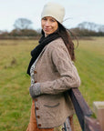 Marina, a white woman with long brown hair is standing sideways, leaning back against a fence in an open field. She is wearing multiple handknitted garments including a knitted cardigan, tank, hat, scarf and mitts.