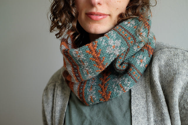 A close up shot of Wandrian, a colourwork cowl knit using hand dyed yarn. It is being modelled by a white woman with mousy coloured curly hair wearing a grey knitted cardigan. The cowl design features abstract patterns of flowers and branches alongside some stripes. The colours used are very autumnal, a lovely teal complimented with a soft grey, a burgundy and a rust orange