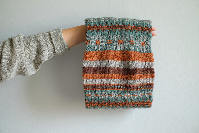 A back shot of Wandrian, a colourwork cowl knit using hand dyed yarn. It is looped over the hand of a white person wearing a grey knitted cardigan and hanging so you can clearly see the colourwork design. The cowl design features abstract patterns of flowers and branches alongside some stripes. The colours used are very autumnal, a lovely teal complimented with a soft grey, a burgundy and a rust orange.