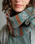 A close up shot of Wandrian, a colourwork cowl knit using hand dyed yarn. It is being modelled by a white woman with mousy coloured curly hair wearing a grey knitted cardigan. The cowl design features abstract patterns of flowers and branches alongside some stripes. The colours used are very autumnal, a lovely teal complimented with a soft grey, a burgundy and a rust orange